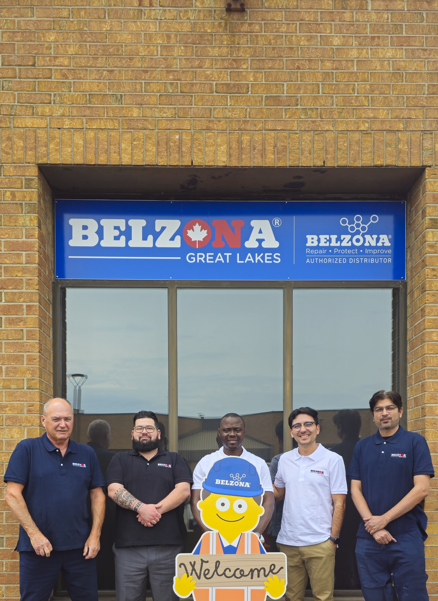 Group of five Belzona Great Lakes team members standing outside their office, smiling, with a colorful cartoon character holding a 'Welcome' sign.